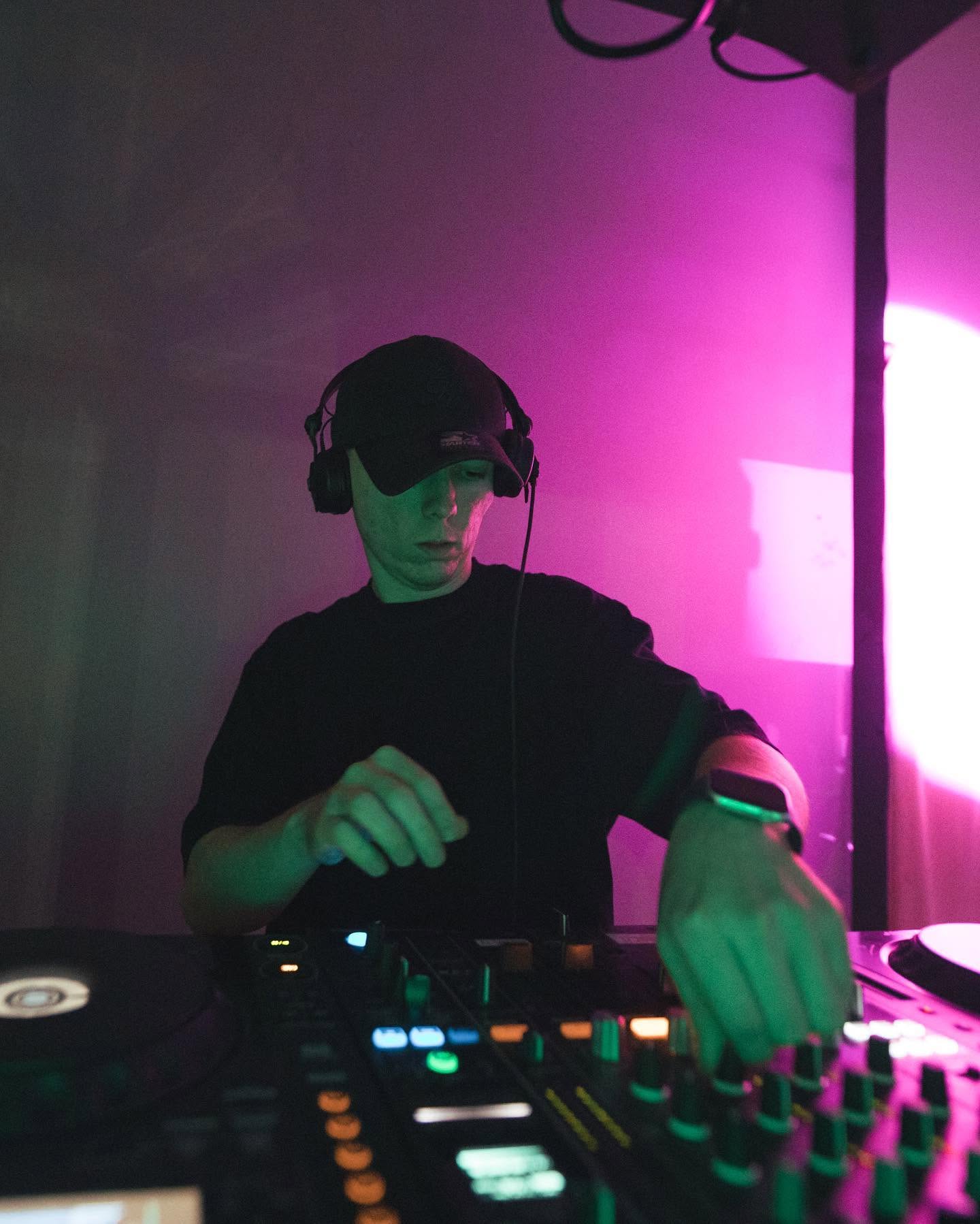 JustG DJing in headphones and a cap under purple and green lighting