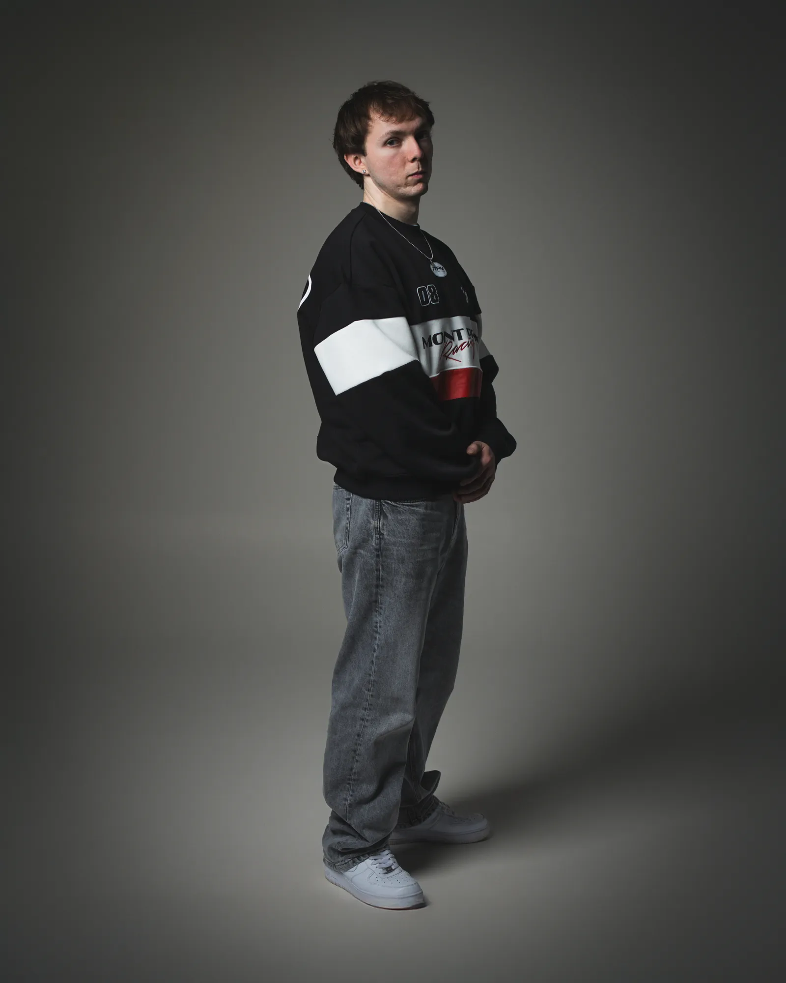 Full-length portrait of JustG in a black, white and red sweatshirt with grey jeans against a grey backdrop