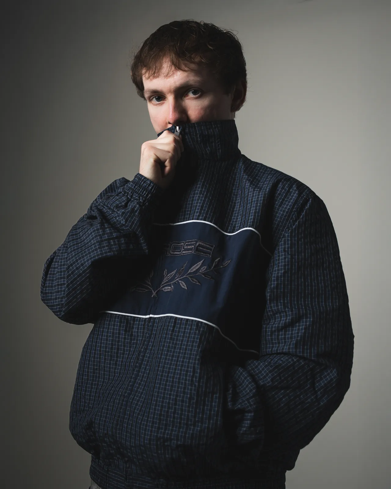 Portrait of JustG in a navy track jacket with the collar pulled up against a grey backdrop